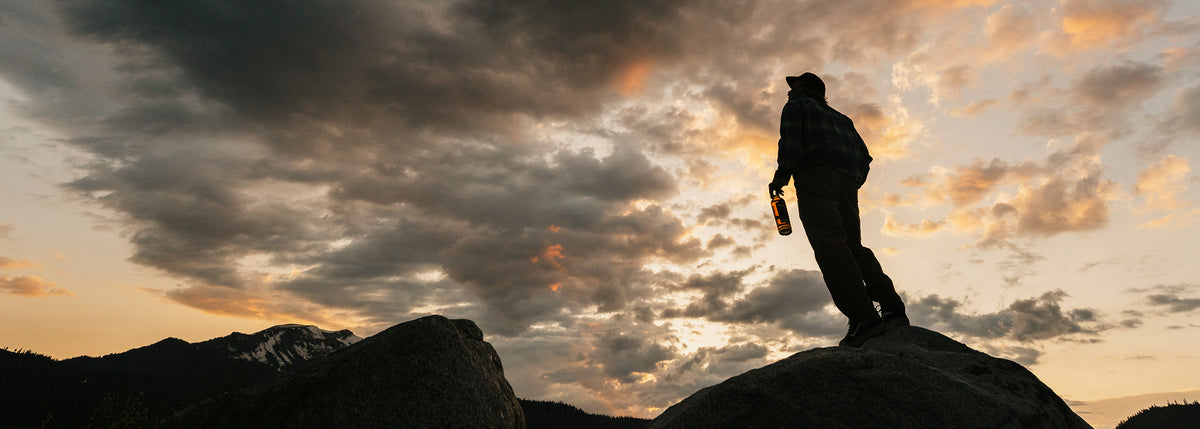 A silhouetted person standing on a rocky outcrop against a dramatic cloudy sky at sunset, holding a bottle. Mountains are visible in the background.