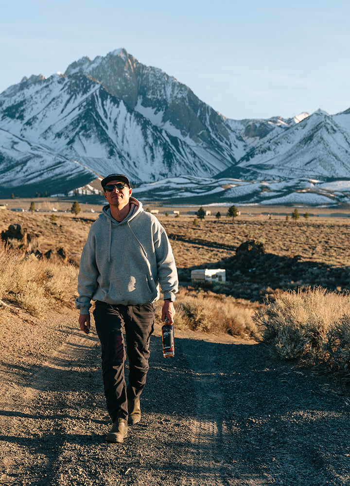 Man walking on a dirt path with snow-capped mountains in the background with a bottle of Timeless Whiskey from Shelter Distilling